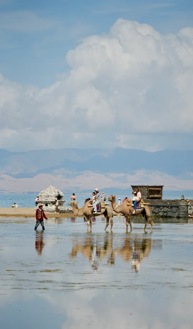 Camels and their riders reflected in shallow water beneath a wide, cloudy sky.